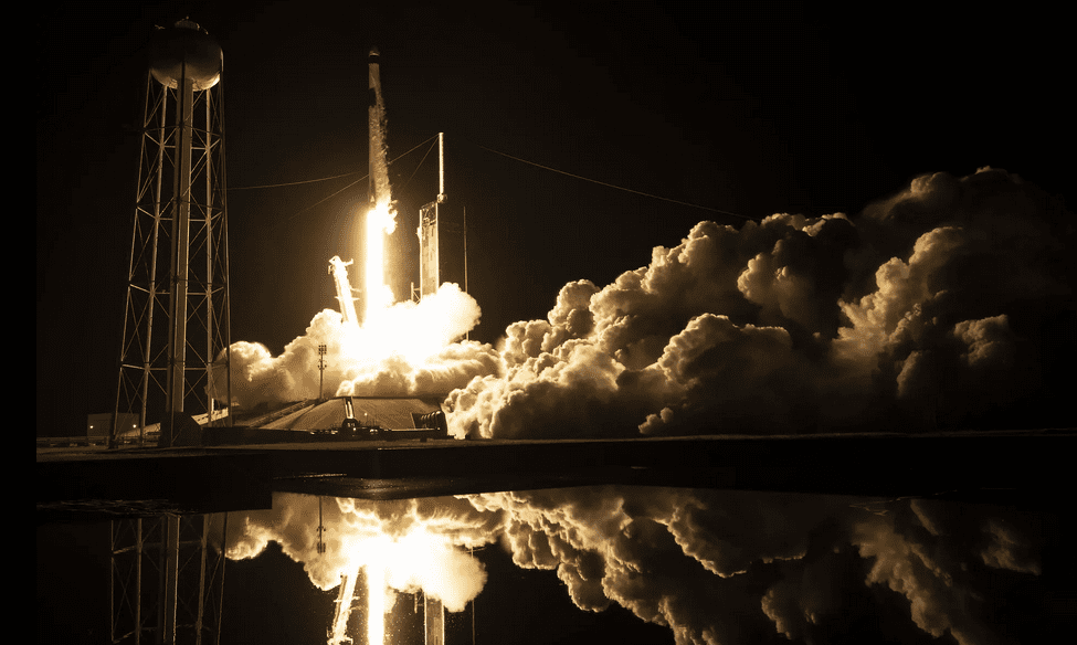 Night rocket launch from Kennedy Space Center, Cape Canaveral, with bright exhaust plumes and reflection.