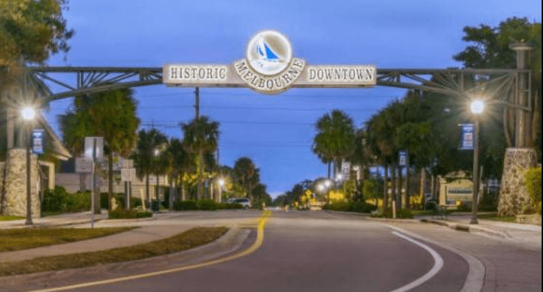 A nighttime photo of the "Historic Downtown Melbourne" arched sign lit up over a street at dusk.